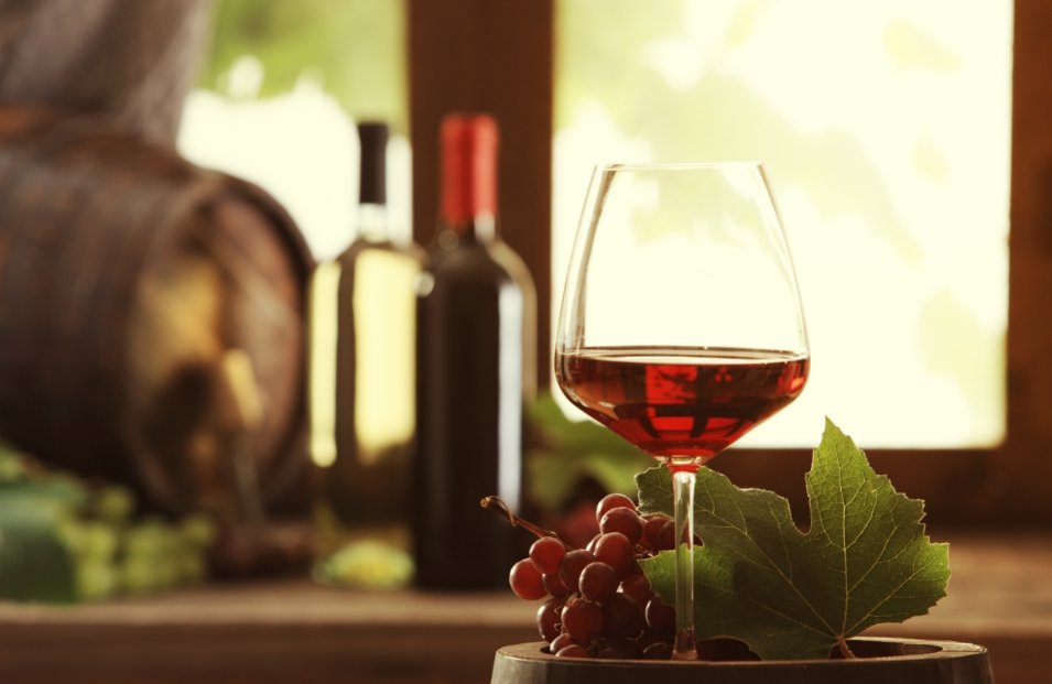 На фотографии изображен бокал с красным вином на фоне бутылки вина и бочки
